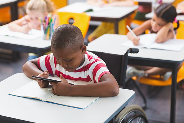 School boy in a wheelchair sitting at a desk using a using digital tablet