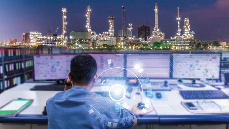 Image of plant worker examining computer monitors and factory in background