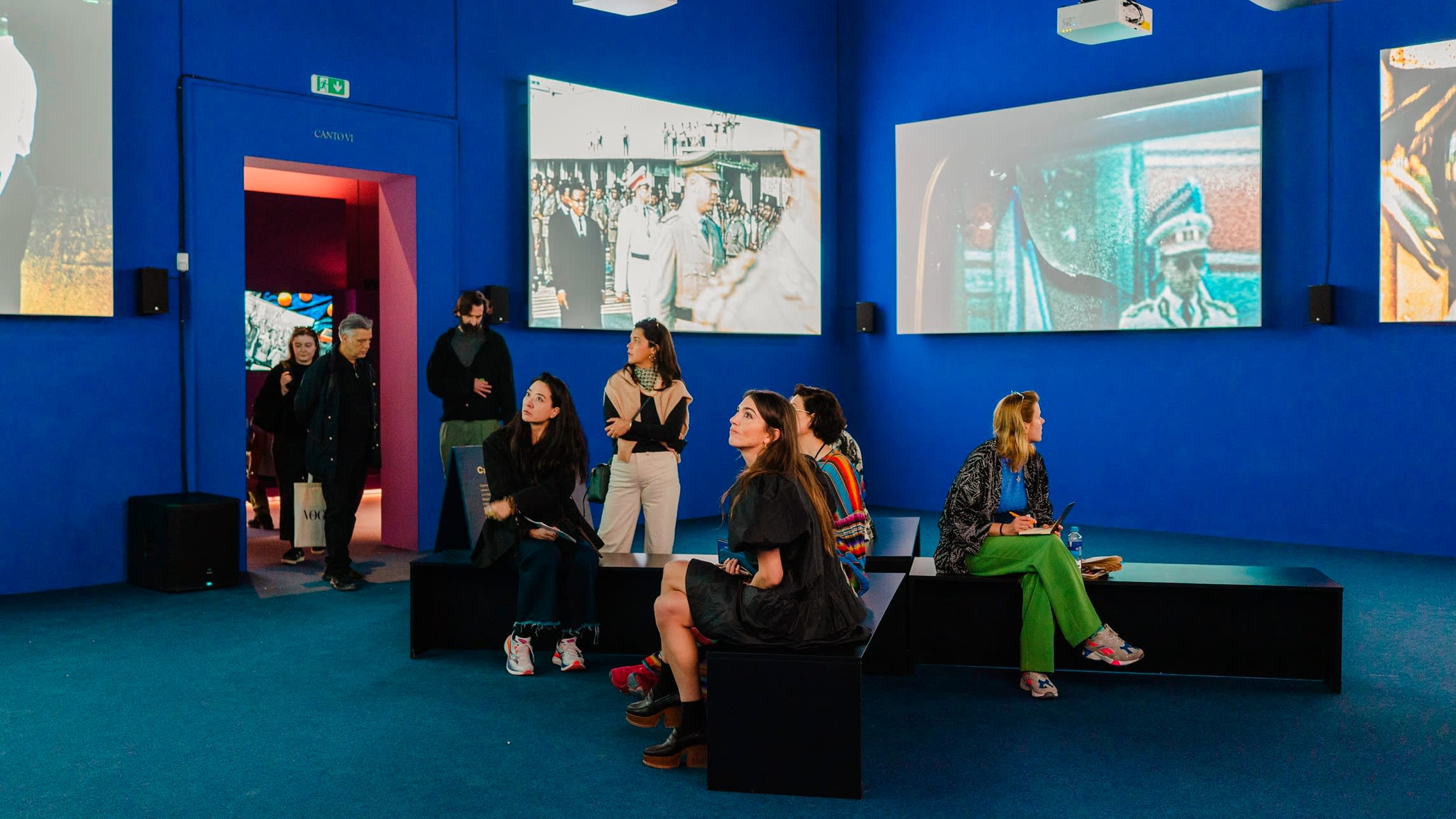 A group of people, mainly young women, inside a blue-walled art gallery with large pictures on its walls