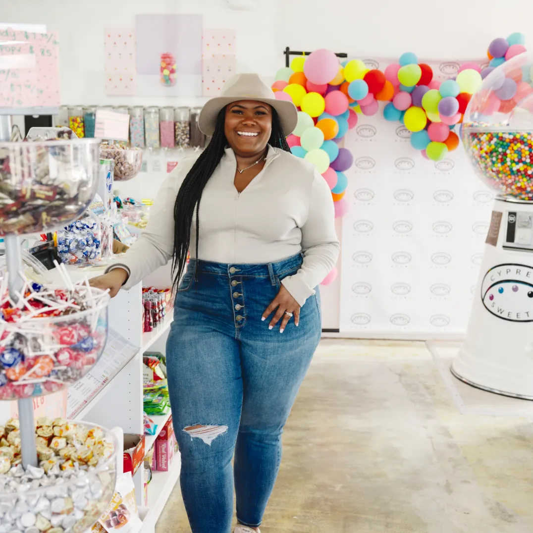 La propietaria de Cypress Sweets posa de pie para una foto en su tienda de golosinas con ropa informal y un sombrero.