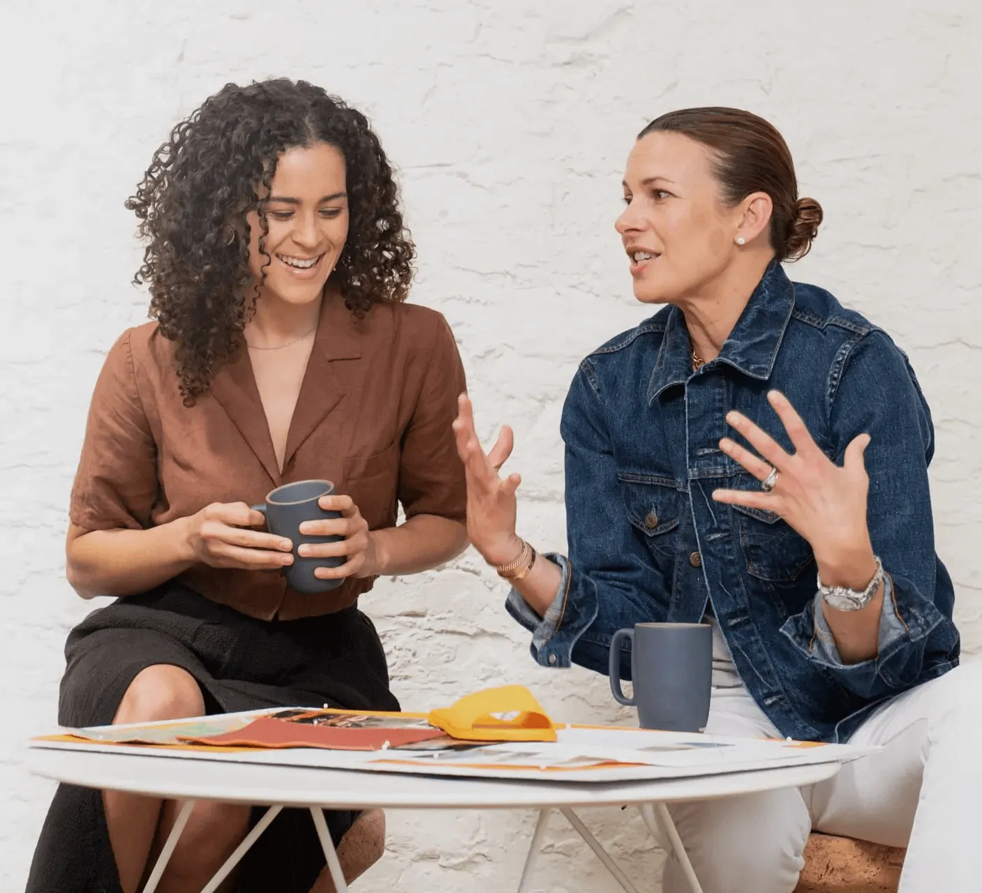 Two women speaking animatedly in front of a white brick wall.