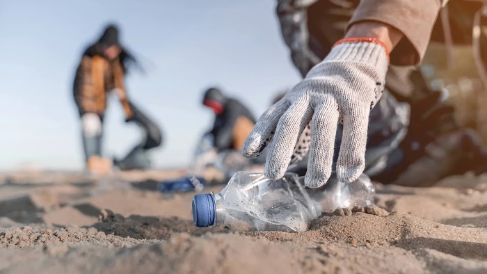 A gloved hand reaches for a plastic water bottle lying on a beach.