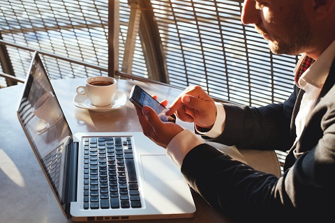 Man sitting in front of a laptop, touching the screen of a smartphone - IMD Business School