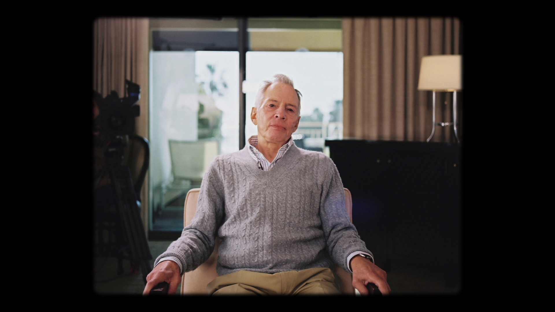 An elderly man in a gray sweater, seated in a room with a glass balcony door visible behind him; still from 'The Jinx - Part Two'