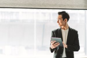 An young businessman holding a digital tablet stands in front of a large window in his modern office.