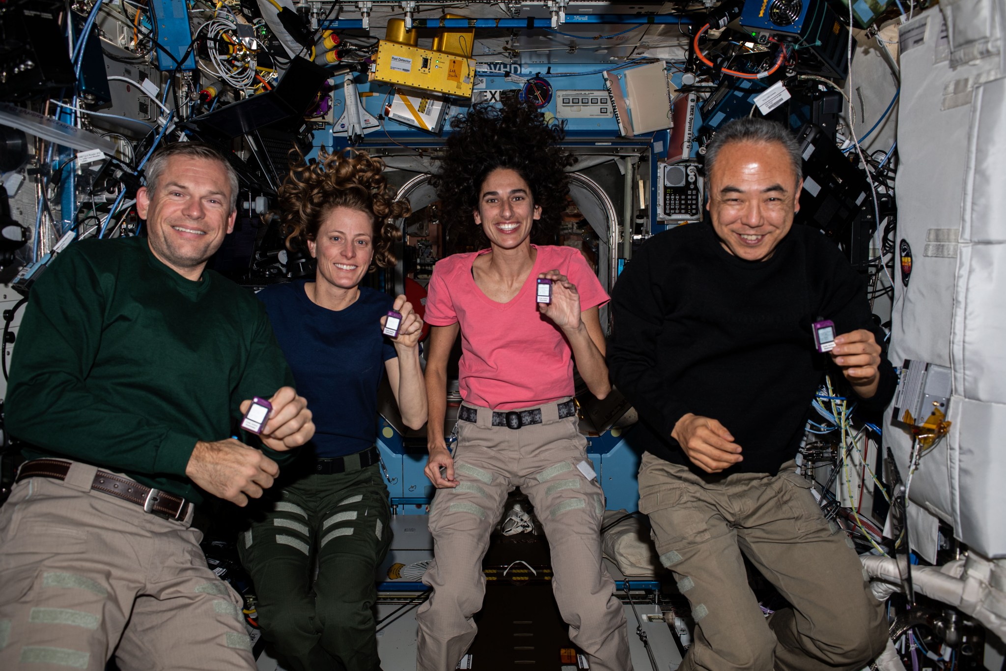 Mogensen is wearing a long-sleeved green shirt and khaki pants, O’Hara a short-sleeved blue shirt and green pants, Moghbeli a short-sleeved pink shirt and khaki pants, and Furukawa a long-sleeved black shirt and khaki pants. All four are smiling at the camera and holding small pager-sized devices that are white on the front and purple around the edges.