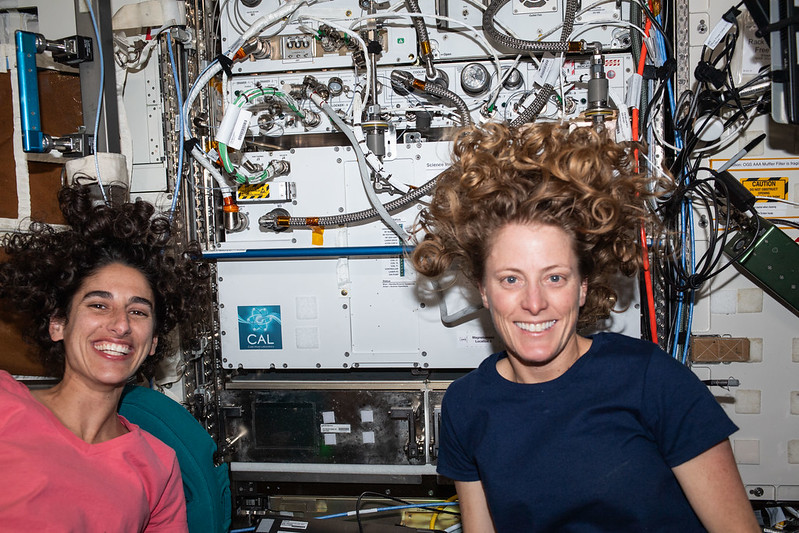 Moghbeli, left, wears a coral-colored t-shirt and O’Hara wears a dark blue t-shirt. Both are smiling at the camera as their curly hair floats around their heads. The white front of the Cold Atom Lab hardware, visible behind them, has multiple hoses and tubes attached to its front.