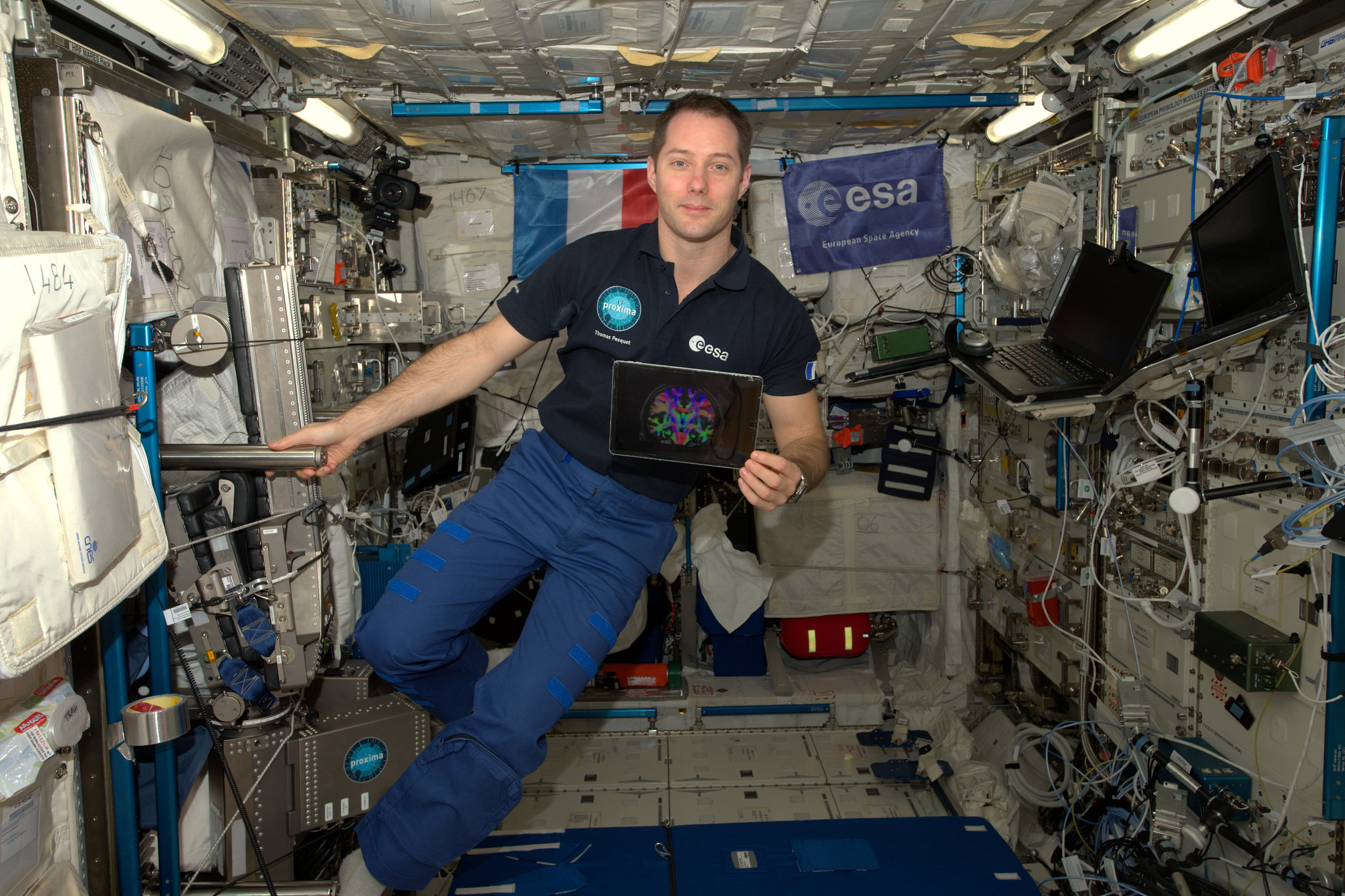 Pesquet viste una camiseta polo de color oscuro con el logotipo de la ESA y pantalones celestes. Mira a la cámara y con la mano derecha se aferra a una barra que se extiende desde una pared de la estación espacial. En su mano izquierda tiene una tableta con una imagen colorida. En la pared de la estación que está detrás de él hay banderas de la ESA y de Francia, y la pared a su izquierda está cubierta por computadoras portátiles, equipos y cables.