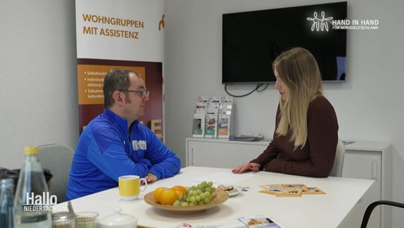 Ein Mann und eine Frau sitzen an einem Tisch, auf dem eine Schale mit Obst steht. An der Wand hängt ein TV. © Screenshot 