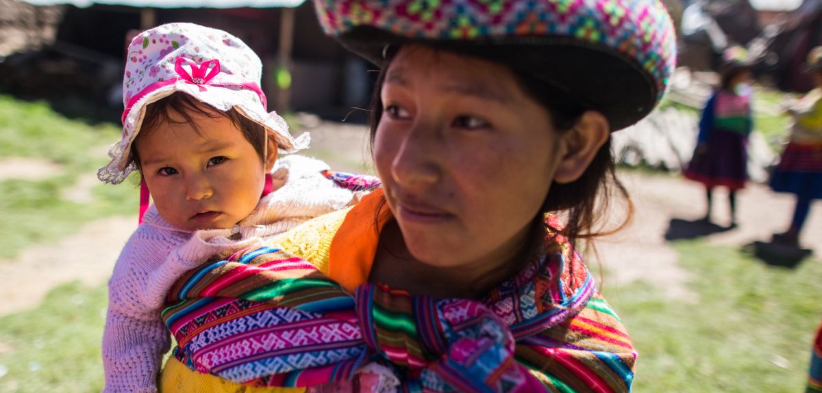 A young Peruvian woman wearing a colourful hat and shawl holds a baby. She is standing outside. Village buildings are visible behind her.