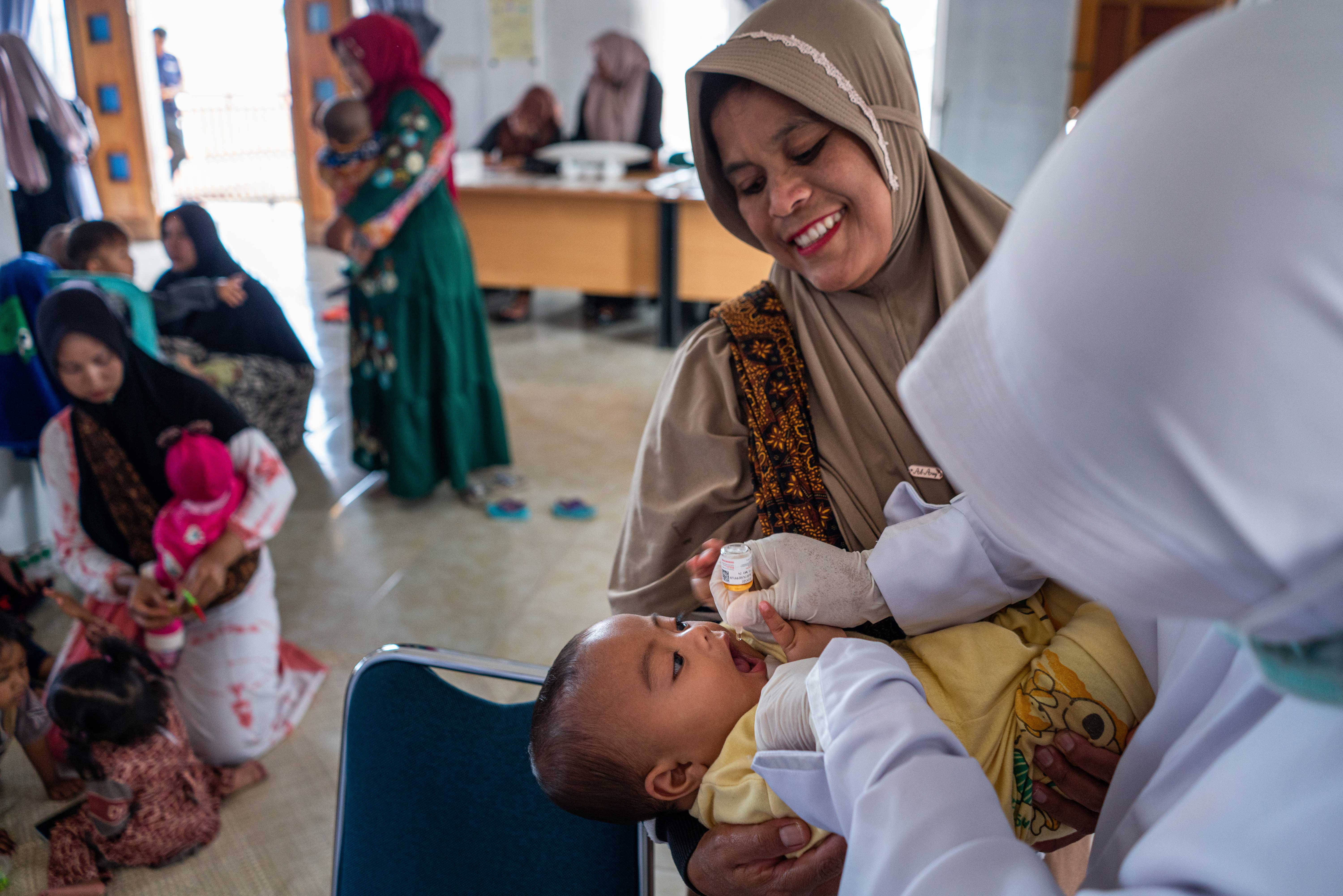 A health professional is administering an oral vaccine or medication to a baby, who is being held by a smiling woman, likely the mother.