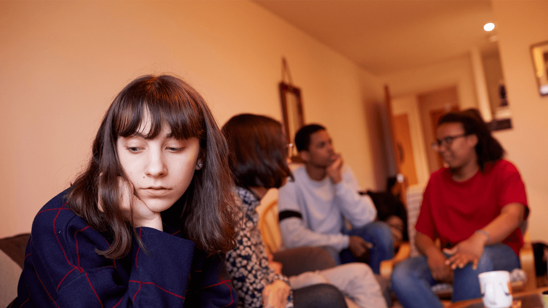 A girl sitting in a living room with her friends. She is looking away as they talk behind her and seems left out of the conversation.